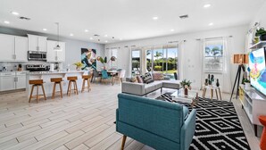 A modern kitchen with white cabinets and a central island, a dining area with a table and chairs, a living room with a sofa and TV, and large windows with a view of greenery. - Modern Family Villa Close To Disney With Private Pool (Kissimmee)