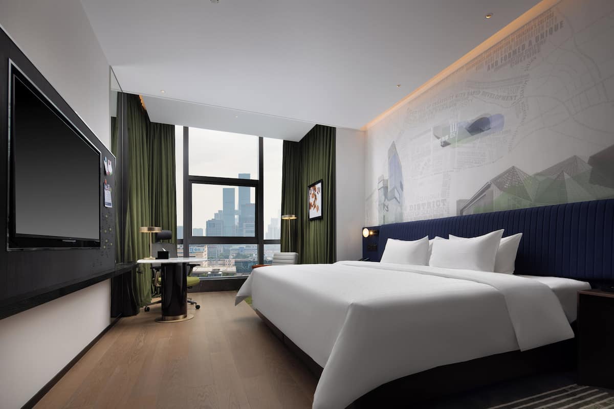 Une chambre d’hôtel moderne dotée d’un grand lit, d’un téléviseur à écran plat, d’un bureau avec une chaise et offrant une vue sur le paysage urbain grâce à une grande fenêtre.