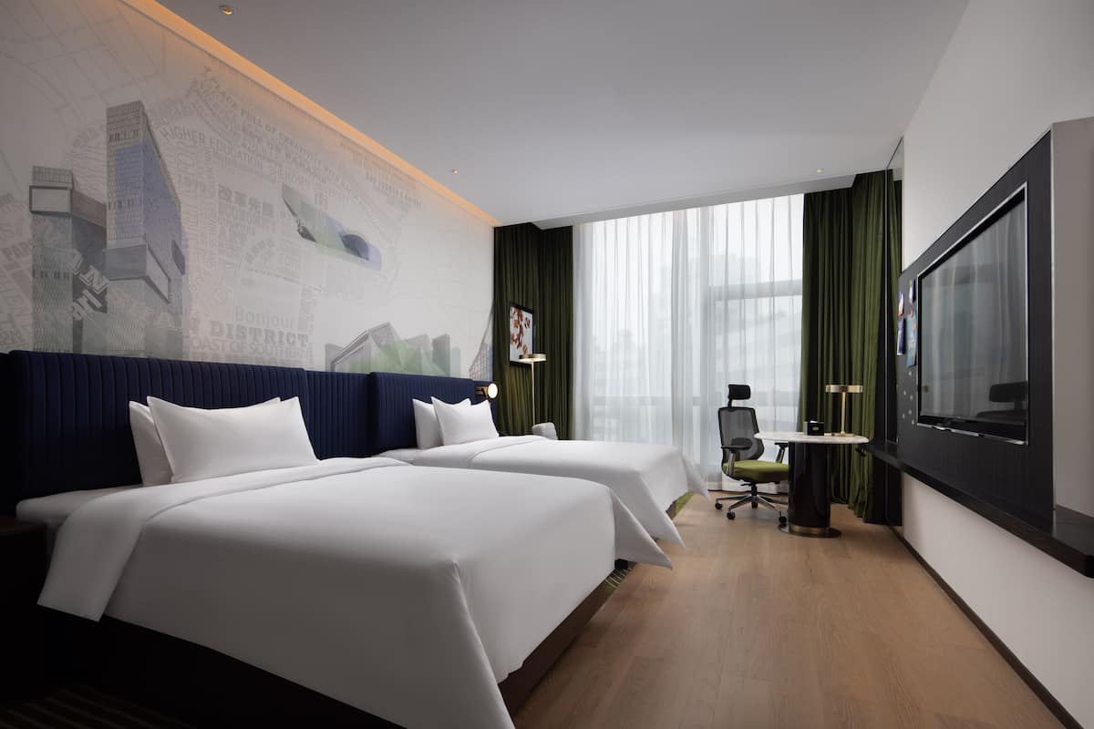 Une chambre d’hôtel avec deux lits, un bureau, une chaise, une grande fenêtre et un mur orné d’une fresque représentant un paysage urbain.