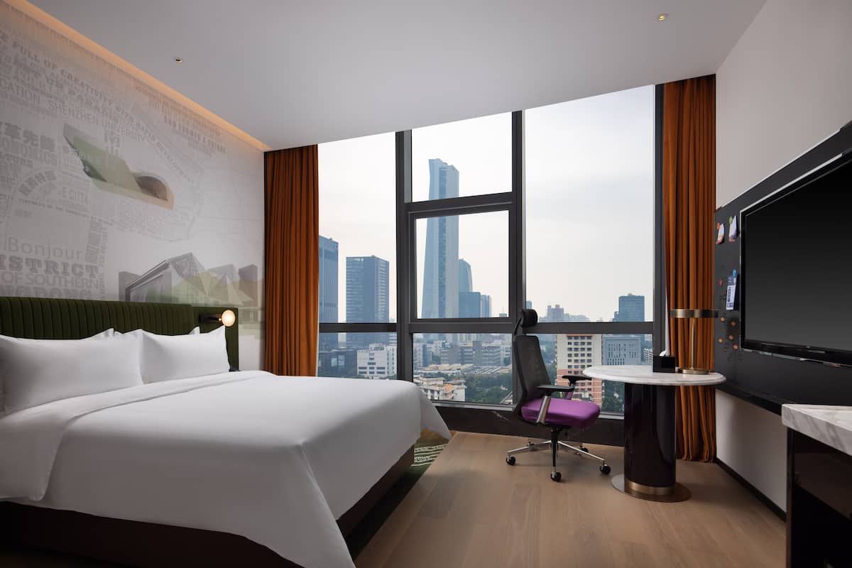 Une chambre d’hôtel dotée d’un grand lit, d’un bureau avec une chaise, d’une télévision et offrant une vue sur le paysage urbain à travers de grandes fenêtres.