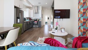 A modern living room with a dining area, a kitchen in the background, and a flat-screen TV mounted on the wall.