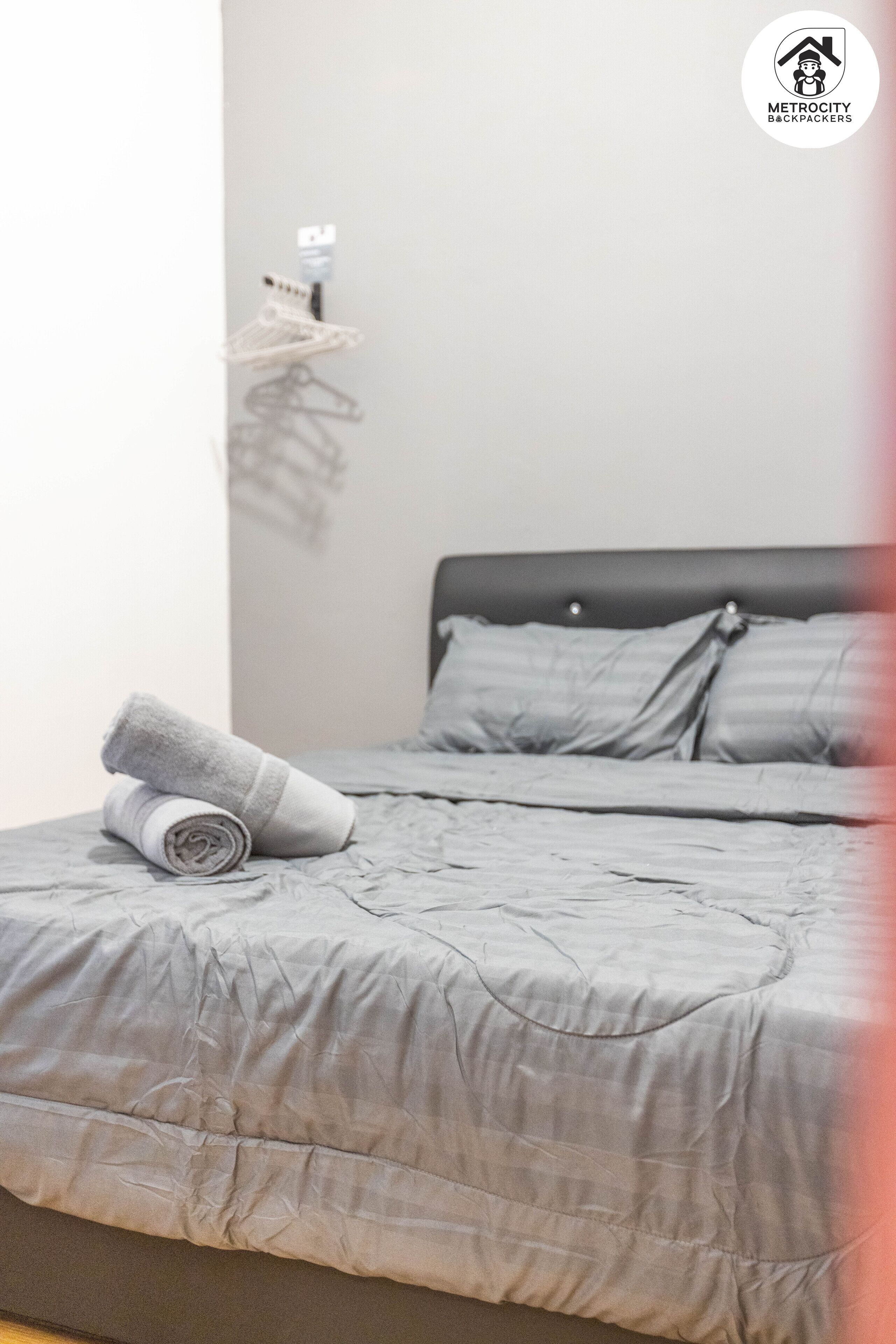 Una cama bien hecha con edredón gris y toalla enrollada, cabecera negra y perchero de pared.