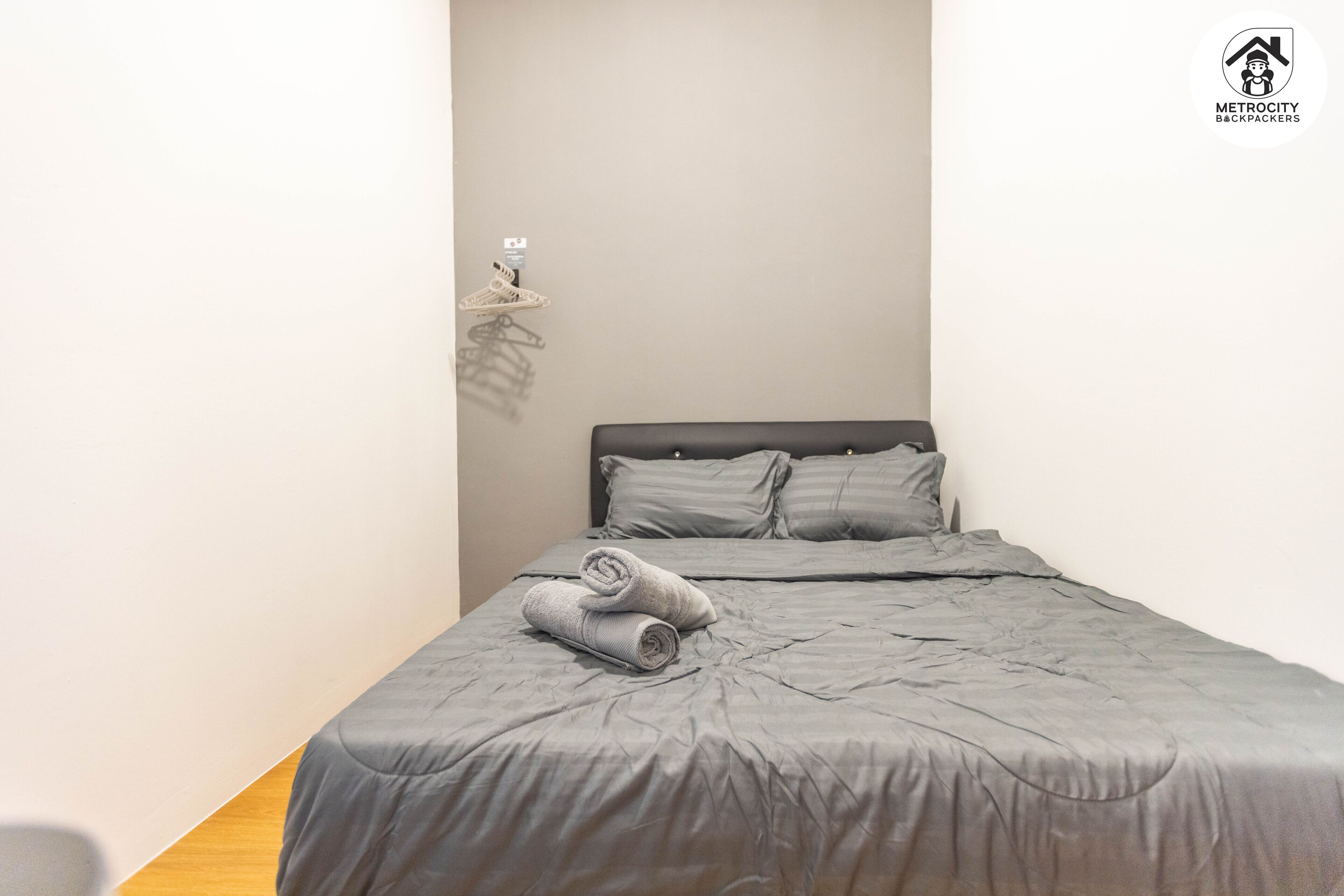 Una cama bien hecha con edredón y almohadas grises, suelo de madera y un perchero para toallas en la pared.