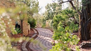 Un chemin sinueux pavé de briques mène à un bâtiment dont la façade est également en briques, et qui est bordé d’un grand arbre.