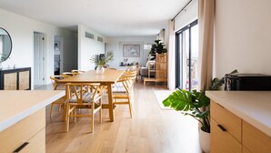 Uma sala de jantar espaçosa com uma longa mesa de madeira e cadeiras, bancada de cozinha e uma grande janela com cortinas.