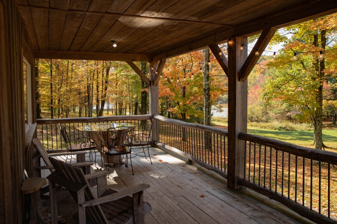 A covered outdoor seating area with a table and chairs, overlooking a forested area with autumn foliage.