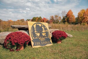 Un letrero de madera con una calavera y árboles, que dice 'THE LODGE', sobre un fondo de follaje otoñal y cielo despejado.