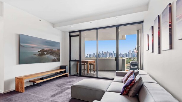 A modern living room with a large window, a sofa, a bench, and a city view. - 2-Bed, 500m From The Beach with Pool, Gym & Sauna (Port Melbourne)