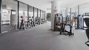 A gym with various exercise machines, a large pillar with a notice, and large windows. - 2-Bed, 500m From The Beach with Pool, Gym & Sauna (Port Melbourne)