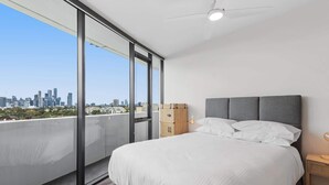 A bedroom with a bed, a nightstand, a suitcase, and a view of a city skyline. - 2-Bed, 500m From The Beach with Pool, Gym & Sauna (Port Melbourne)