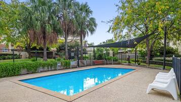 A rectangular swimming pool with a tiled edge, surrounded by a fence, and shaded by a canopy.