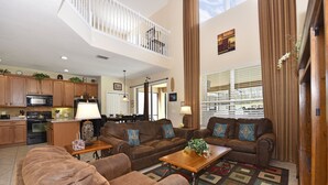 A spacious living room with a brown leather sofa set, a wooden coffee table, and a patterned rug. A kitchen with wooden cabinets and a microwave is visible in the background. - Fs3901ha - 6 Bedroom Villa In Watersong, Sleeps Up To 12, Just 9 Miles To Disney (Davenport)