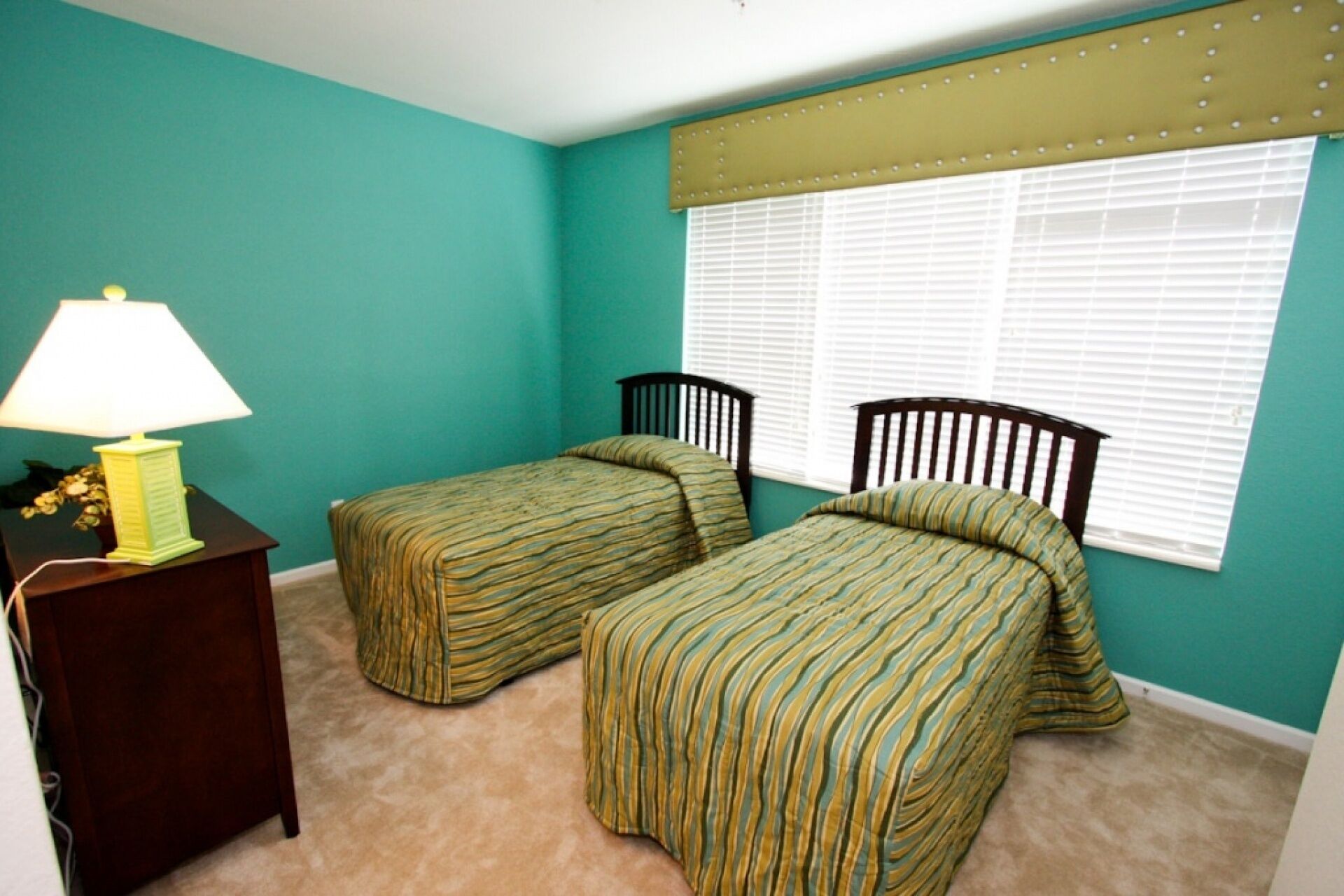 A bedroom with teal walls, two single beds with striped bedding, a nightstand with a lamp, and a window with blinds.