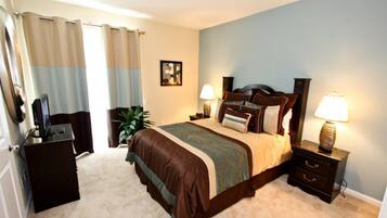A bedroom with a large bed, two bedside tables with lamps, a dresser, a television, and a plant.