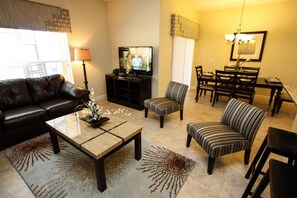 A living room with a leather sofa, a coffee table with a floral centerpiece, a TV, and a dining area with chairs and a table. - It1107 - 4 Townhome in Paradise Palms Resort, Sleeps Up To 12, Just 6 Miles To Disney (Kissimmee)