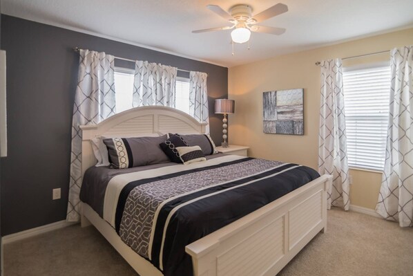 A bedroom with a bed, curtains, a ceiling fan, a nightstand with a lamp, and a painting on the wall.