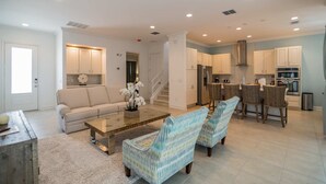 A spacious living room with a sofa, coffee table, and patterned chairs. A kitchen with white cabinets and a dining area with wicker chairs is visible in the background.