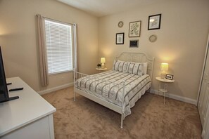 Een slaapkamer met een bed met wit metalen frame, een beige vloerkleed, een raam met zonwering, twee nachtkastjes met lampen en wanddecoraties.