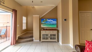 Ein Wohnzimmer mit Fernseher auf einem weißen Schrank, einer Treppe zu einem Balkon und einer Tür rechts.