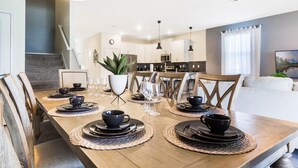 Un coin repas avec une table en bois dressée pour six personnes, entourée de chaises. On aperçoit, au fond, une cuisine équipée d’appareils électroménagers modernes.