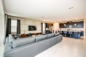 A modern living room with a large grey sectional sofa, a flat-screen TV mounted on the wall, and a kitchen area with dark cabinets and a central island with bar stools. - Fs9601ha - Solara Resort - 9 Bed 6 Baths Villa (Kissimmee)