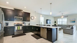 Une cuisine moderne avec des meubles de couleur sombre, des appareils en acier inoxydable, un grand îlot central et un canapé gris dans l’espace salon.