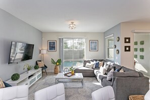 A modern living room with a flat-screen TV, a sofa set, a coffee table, and a large window with a view of a pool. - Fs9665ha - Champions Gate Resort - 4 Bed 3 Baths Townhouse (Davenport)