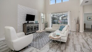 A spacious living room with a white sofa, a white armchair, a TV on a textured wall, and a large window. - Fs9741ha - Storey Lake Resort - 5 Bed 5 Baths Villa (Kissimmee)
