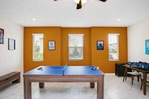 A room with a ping pong table, windows with blinds, a foosball table, and framed pictures on the walls. - Fs9826ha - Storey Lake Resort - 9 Bed 5 Baths Villa (Kissimmee)