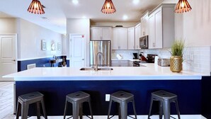 Une cuisine moderne dotée d’un grand îlot central, d’appareils électroménagers en acier inoxydable et de luminaires suspendus.
