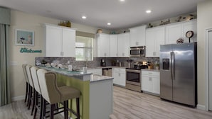 A modern kitchen with white cabinets, stainless steel appliances, a central island with bar stools, and a "Welcome" sign on the wall. - Grh9266ha - Champions Gate Resort - 6 Bed 6 Baths Villa (Davenport)