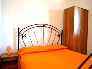 Une chambre à coucher avec un lit en métal, une couvre-lit orange, une armoire en bois et une table de chevet avec une lampe.