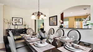 Un espace repas avec une longue table dressée pour six personnes, agrémenté d’un lustre, d’œuvres d’art encadrées et offrant une vue sur la cuisine.