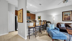 A modern kitchen with a refrigerator, microwave, and stove. There are wooden cabinets and a bar area with stools. A blue ottoman and a brown couch are in the living area. - Grh9352ha - Paradise Palms Resort - 4 Bed 3 Baths Townhome (Kissimmee)