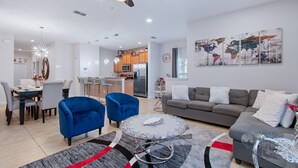 A modern living room with a grey sofa, blue armchairs, and a coffee table. There is a kitchen in the background with wooden cabinets and a stainless steel refrigerator. - Grh10334ha - Paradise Palms Resort - 5 Bed 4 Baths Townhome (Kissimmee)