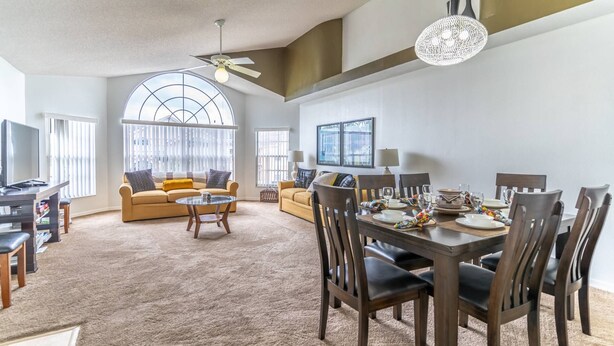 A spacious living room with a large window, a dining area with a table set for four, and a ceiling fan. - It9921 - 4 Condo in Royal Palm Bay Resort, Sleeps Up To 8, Just 5 Miles To Disney (Kissimmee)