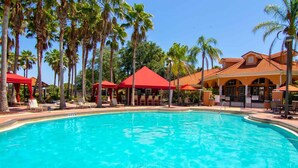 Ein Swimmingpool mit Liegestühlen und Sonnenschirmen, umgeben von Palmen und einem Gebäude mit rotem Dach.