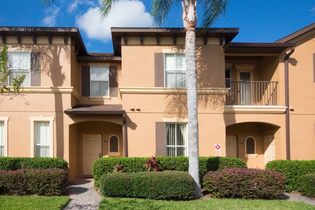 A two-story residential building with a beige exterior, multiple windows with shutters, and a small balcony.