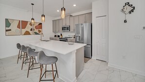 A modern kitchen with a central island, bar stools, pendant lighting, stainless steel appliances, and abstract wall art. - Fs11330ha - Storey Lake Resort - 4 Bed 3 Baths Townhouse (Kissimmee)