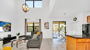 A spacious living room with a grey sofa, a yellow armchair, and a glass coffee table. A kitchen area with wooden cabinets and a large island is visible. - Fs11371ha - Veranda Palms Resort - 6 Bed 4 Baths Villa (Kissimmee)