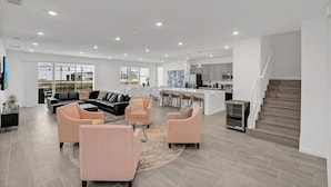 A spacious living area with a black sectional sofa, two pink armchairs, and a coffee table. A kitchen with a bar and a staircase is visible in the background. - Fs11377ha - Veranda Palms Resort - 15 Bed 14.5 Baths Villa (Kissimmee)