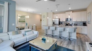 A modern living room with a white sofa, a glass coffee table, and a dining area with a white table and chairs. The kitchen features white cabinets, a stainless steel refrigerator, and a bar with stools. - Fs11384ha - Storey Lake Resort - 4 Bed 3 Baths Townhouse (Kissimmee)