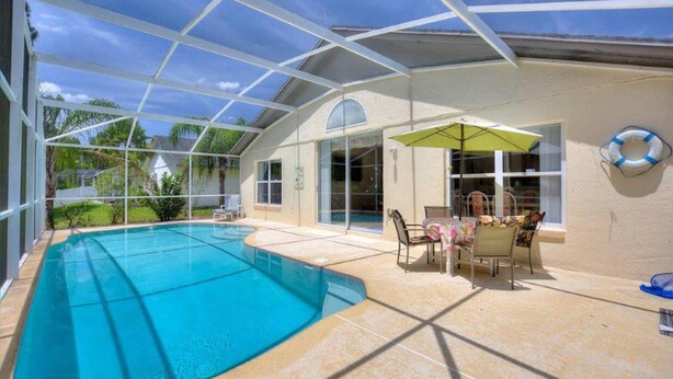 Una piscina al aire libre con cerramiento de vidrio, un área de patio con sillas y una mesa, y un flotador de salvamento en la pared.
