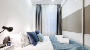 Habitación de hotel moderna con una cama bien hecha, una lámpara de noche, una mesita con lámpara y vistas a la ciudad a través de amplios ventanales.