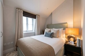 A hotel room with a bed, a nightstand with a lamp, and a window with curtains. - Mayfair`s Finest: Modern 2 bedroom apartment! (Greater London)