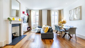 Una sala de estar moderna con un sofá, una mesa de comedor y sillas. Hay una chimenea con un televisor encima, y un espejo grande en la pared.