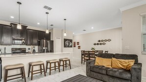 A modern kitchen with dark cabinets, stainless steel appliances, and pendant lighting. There is a dining area with a gray sofa and a coffee table.