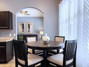A dining area with a round table, four chairs, a ceiling fan, two framed artworks, a small table with a lamp and flowers, and a window with sheer curtains. - Fs14388ha - Dahlia Reserve at Crescent Lakes - 4 Bed 3 Baths Villa (Kissimmee)