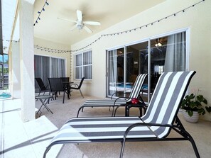 A covered patio area with striped lounge chairs, a table, and chairs, and a view of a swimming pool through large glass doors. - Fs14388ha - Dahlia Reserve at Crescent Lakes - 4 Bed 3 Baths Villa (Kissimmee)