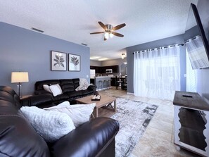 A modern living room with a black leather sofa, a wooden coffee table, and a ceiling fan. - Fs14388ha - Dahlia Reserve at Crescent Lakes - 4 Bed 3 Baths Villa (Kissimmee)